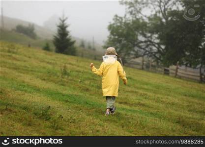 Cute little girl in yellow raincoat is walking in foggy mountains. Bad weather, summer tropical storm, autumn fashion concept. back view.. Cute little girl in yellow raincoat is walking in foggy mountains. Bad weather, summer tropical storm, autumn fashion concept. back view