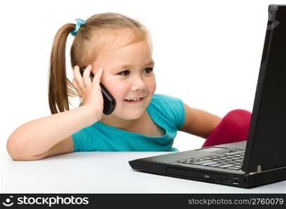 Cute little girl is sitting at table with her black laptop and talking to a cell phone, isolated over white