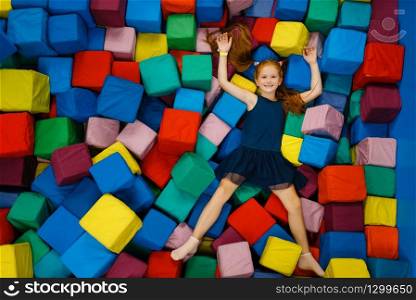 Cute little girl lying in soft cubes, playground in entertainment center. Play area indoors, playroom