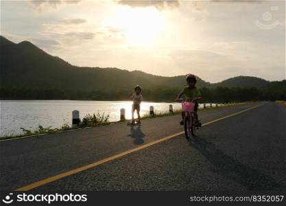 Cute little girl riding a bicycle and her sister riding a scooter on a lake road at sunset. Happy sisters doing outdoor activities together. Healthy Summer Activities for Kids.