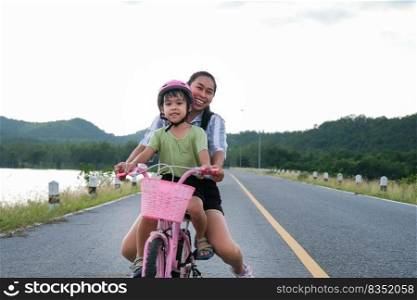 Cute little girl riding a bicycle with her mother on a lake road at sunset. Happy family doing outdoor activities together. Healthy Summer Activities for Kids.