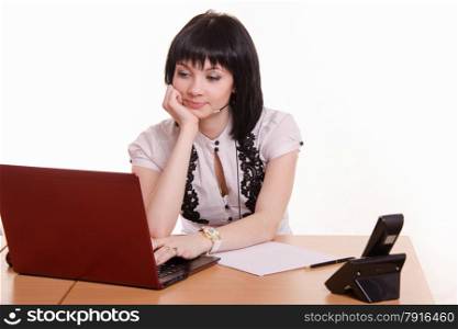 Cute little girl sitting at a table in the call center with laptop in white blouse propped his head on his hand. Office employee call-center resting on hand looking at the monitor