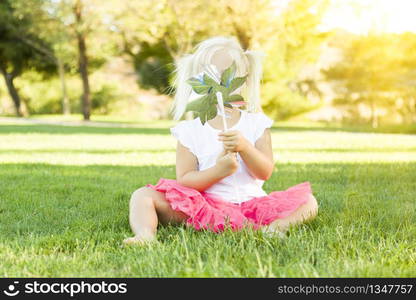 Cute Little Girl Sitting In Grass Blowing On Pinwheel Toy.