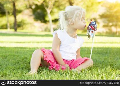 Cute Little Girl Sitting In Grass Blowing On Pinwheel Toy.