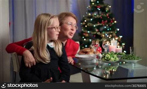 Cute little girl sitting on grandmother&acute;s lap and telling story on Christmas Day in living room at home over Christmas tree background.