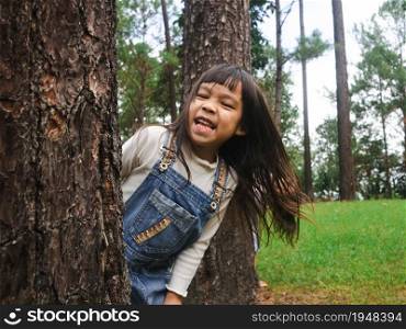 Cute little girl standing behind a big tree and smiling at the camera. Happy child playing outdoors. Family spent time together.