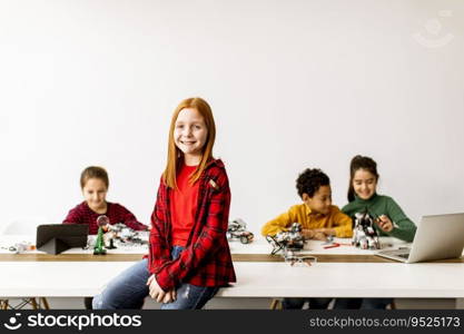 Cute little girl standing in front of  group of kids programming electric toys and robots at robotics classroom