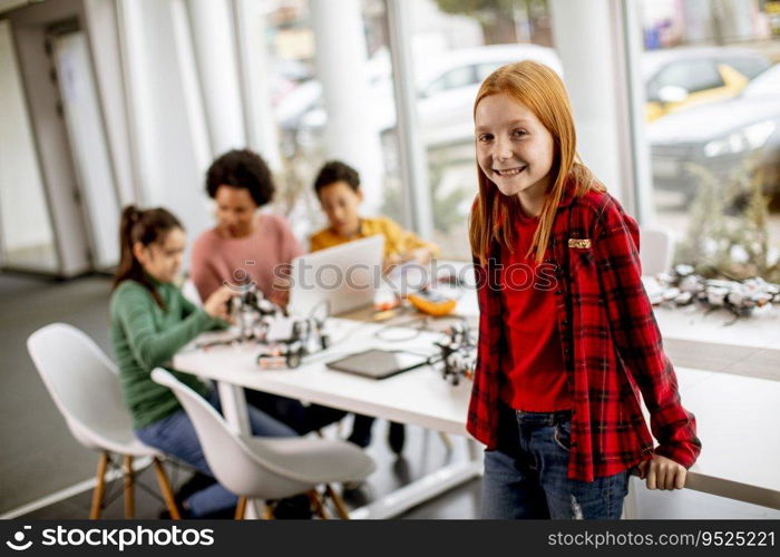 Cute little girl standing in front of  group of kids programming electric toys and robots at robotics classroom