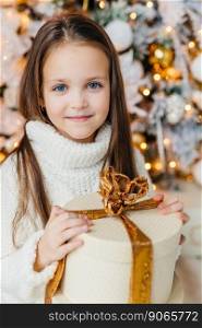 Cute little girl standing near Christmas tree and holding round gift box decorated with golden ribbon. . Girl with gift box