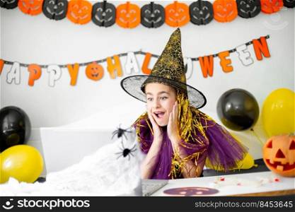 Cute little girl wearing witch hat sitting behind a table in Halloween theme decorated room. Halloween party concept. Halloween with safety measures from Covid-19. Cute little girl wearing witch hat sitting behind a table in Halloween theme decorated room. Halloween party concept.