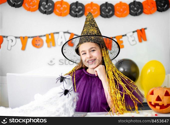 Cute little girl wearing witch hat sitting behind a table in Halloween theme decorated room. Halloween party concept. Halloween with safety measures from Covid-19. Cute little girl wearing witch hat sitting behind a table in Halloween theme decorated room. Halloween party concept.