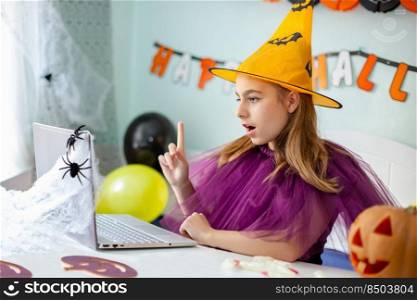 Cute little girl wearing witch hat sitting behind a table in Halloween theme decorated room. Halloween party concept. Preparation for the celebration, internet communication.. Cute girl wearing witch hat sitting behind table Halloween theme decorated room. Preparation for the celebration.