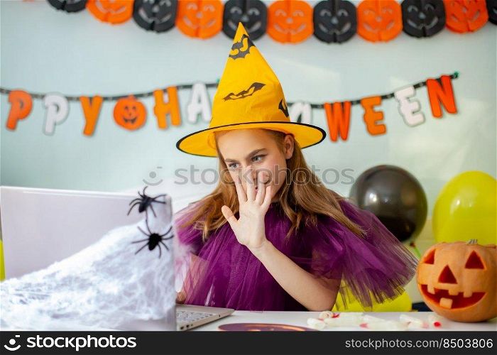 Cute little girl wearing witch hat sitting behind a table in Halloween theme decorated room. Halloween party concept. Preparation for the celebration, internet communication.. Cute little girl wearing witch hat sitting behind table Halloween theme decorated room. Preparation for the celebration.