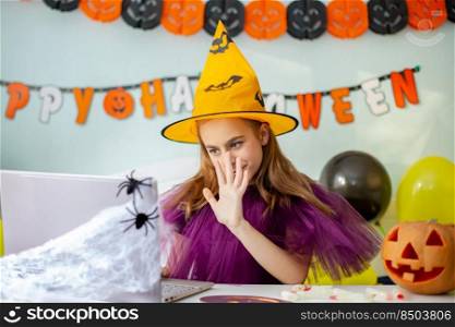 Cute little girl wearing witch hat sitting behind a table in Halloween theme decorated room. Halloween party concept. Preparation for the celebration, internet communication.. Cute little girl wearing witch hat sitting behind table Halloween theme decorated room. Preparation for the celebration.