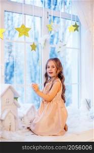 Cute little girl with curly hair at the window in anticipation of Christmas and New Year&rsquo;s magic. Funny emotions in winter day.. Cute little girl with curly hair at the window in anticipation of Christmas and New Year&rsquo;s magic