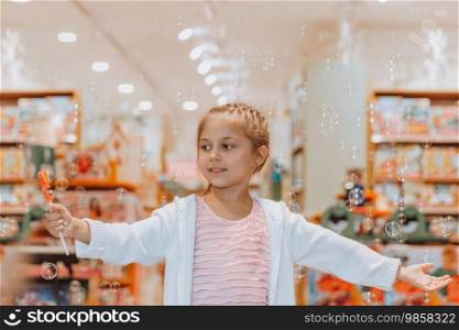 Cute little girl with pleasure blowing soap bubbles in big beautiful toy store. Happy child having fun in shopping mall. Happiness in her eyes.. Little Girl Blowing Soap Bubbles