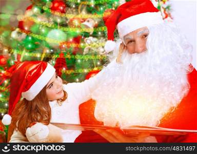 Cute little girl with Santa Claus sitting near Christmas tree and reading Magic xmas story, fairy tale, cozy festive eve at home