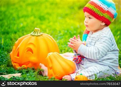 Cute little kid sitting on green grass field wearing funny colorful hat and playing with pumpkins with smiling carved faces, happy Halloween holiday