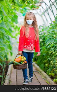 Cute litttle girl wearing a mask for prevent virus with big busket full of vegetables in greenhouse. Harvesting time. Cute little girl collects crop cucumbers and tomatos in greenhouse