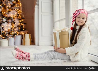 Cute lovely girl with present, sits near window in living room, looks delightfully into camera, admires wonderful beautiful decorated fir tree, has good mood and holidays. New Year concept