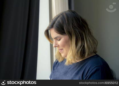 Cute lovely shy smiling young woman standing near the window.