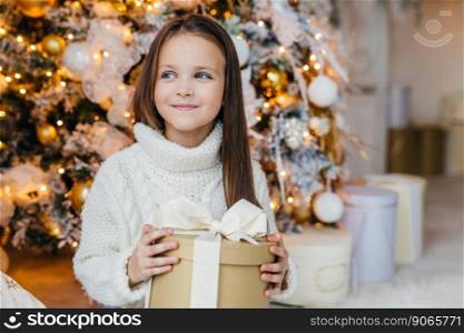 Cute lovely small kid with charming appearance, glad to recieve Christmas gift, looks aside with happy expression, has intrgue what present is, poses near beautiful decorated fir tree.