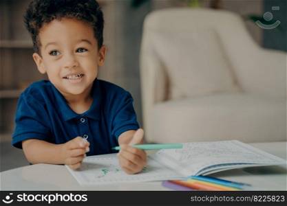 Cute mixed race small boy in polo tshirt painting coloring book with felt tip pens, sitting by table in living room against blurred background with armchair. Children leisure time at home concept. Cute afro american small boy in dark blue polo tshirt painting coloring book at home
