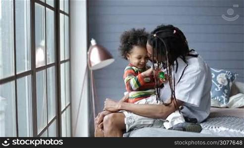 Cute mixed race son sitting on father&acute;s lap and playing with helicopter toy. Stylish african american father with dreadlocks sitting on the bed and holding his little toddler boy on the lap, expressing his love with gentle kisses at domestic interior