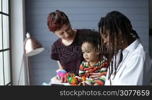 Cute mixed race toddler boy having fun with parents in bedroom at home. Interracial family lounging together. Handsome african father with dreadlocks and cheerful caucasian mother playing together with son with toys while child sitting on dad&acute;s lap.