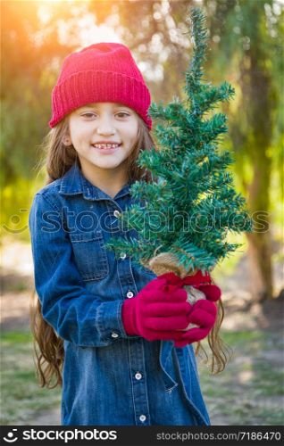 Cute Mixed Race Young Girl Wearing Red Knit Cap and Mittens Holding Small Christmas Tree Outdoors.