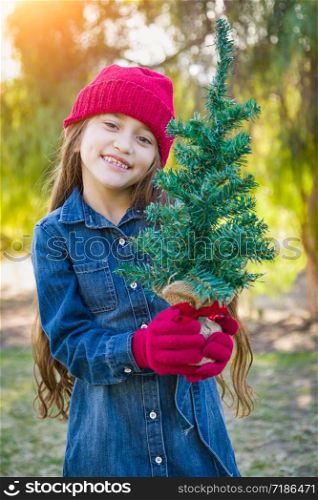Cute Mixed Race Young Girl Wearing Red Knit Cap and Mittens Holding Small Christmas Tree Outdoors.