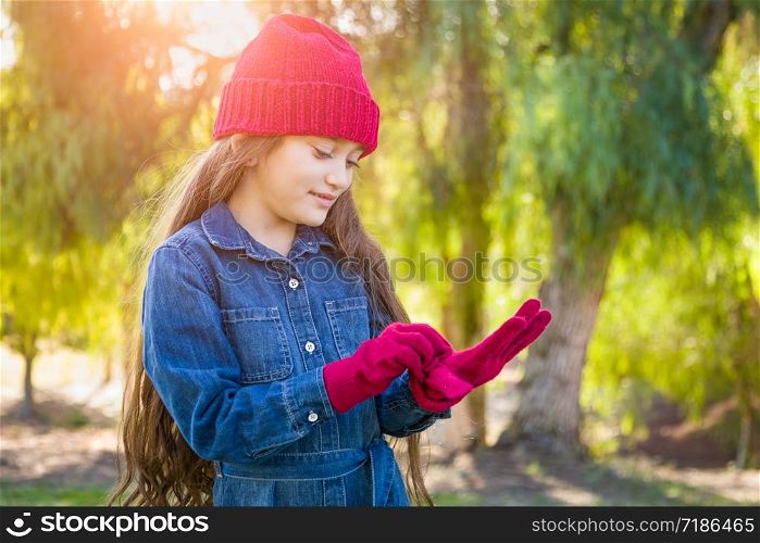 Cute Mixed Race Young Girl Wearing Red Knit Cap Putting On Mittens Outdoors.