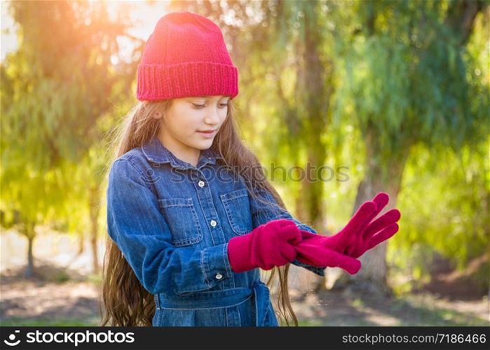 Cute Mixed Race Young Girl Wearing Red Knit Cap Putting On Mittens Outdoors.