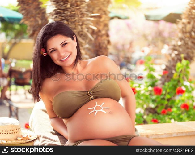 Cute pregnant female on the beach, tanning with drawing sun on the belly, using sunblock for skin protection, healthy lifestyle, enjoying active summer vacation