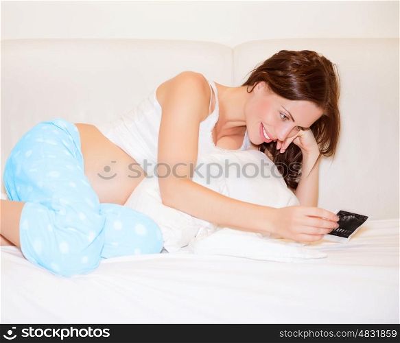Cute pregnant woman lying down on bed at home and enjoying photo of ultrasound of her baby, happy parenthood concept&#xA;