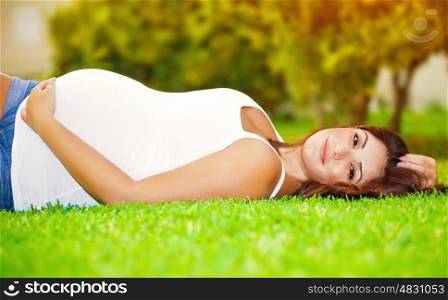 Cute pregnant woman lying down on fresh green grass on the backyard, happy parenthood, healthy pregnancy, spring season