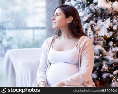 Cute pregnant woman sitting near beautiful decorated Christmas tree at home, looking in window on snowfall, happy pregnancy time
