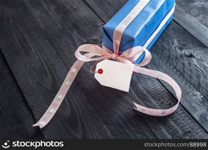 Cute present wrapped in blue paper and tied with pink ribbon and bow, with a blank tag attached to it, on an old wooden table.