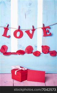 Cute red gift box with a blank message card near it and the word love written in red paper letters tied to a string, on a blue wooden fence.