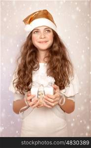 Cute Santa helper, pretty teen girl wearing festive hat and holding in hands gift box, snow fall, looking up, Christmas party concept