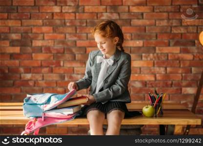 Cute schoolgirl puts textbook into the schoolbag, side view. Female pupil with backpack, desk with textbooks, apple and globe on backgrpund, young girl in the school. Cute schoolgirl puts textbook into the schoolbag