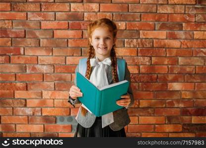 Cute schoolgirl with schoolbag holds an open textbook, brick wall on background. Adorable female pupil with backpack and books poses in the school. Cute schoolgirl with schoolbag holds textbooks
