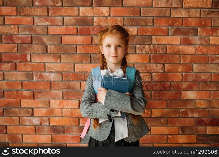 Cute schoolgirl with schoolbag holds textbooks, brick wall on background. Adorable female pupil with backpack and books poses in the school