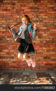 Cute schoolgirl with schoolbag jumps at the brick wall. Playful female pupil with backpack poses in the school