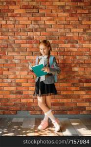 Cute schoolgirl with schoolbag reading a textbook, brick wall on background. Adorable female pupil with backpack and book poses in the school