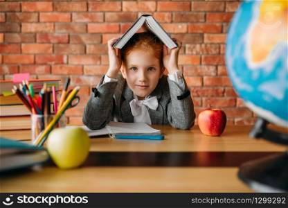 Cute schoolgirl with textbook on her head sitting at the table. Female pupil reading a book at the desk, young girl doing homework