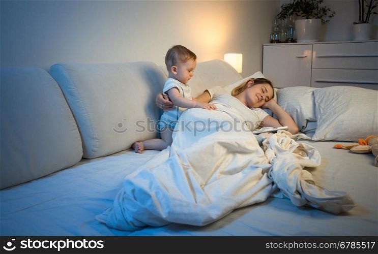 Cute sleepless baby boy waking up his mother sleeping in bed