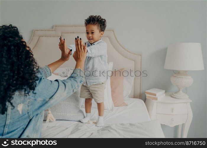Cute small afro american kid playing engaging games on big bed in light themed bedroom with his caring curly mother, holding their hands together. Loving motherhood and child care concept. Cute happy small afro american kid playing with his mom in bedroom at home