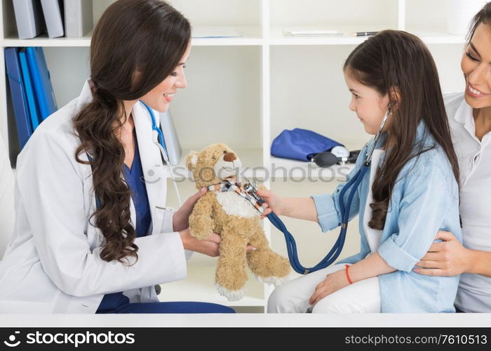 Cute small child patient pretending to be doctor, holding teddy bear toy at meeting with general practitioner. Friendly nurse showing how stethoscope works to smiling little girl at checkup in clinic.. Child patient with general practitioner