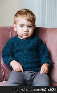Cute small child with light hair and blue eyes, wears sweater and trousers, has innocent expression, looks directly into camera. Adorable little kid has confident look, sits on comfortable armchair